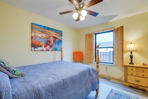 Quarto com cama, ventoinha no teto, janela com cortinas, mesa de cabeceira e um quadro na parede.