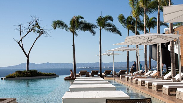 A poolside area with lounge chairs and umbrellas, overlooking a body of water with mountains in the distance.
