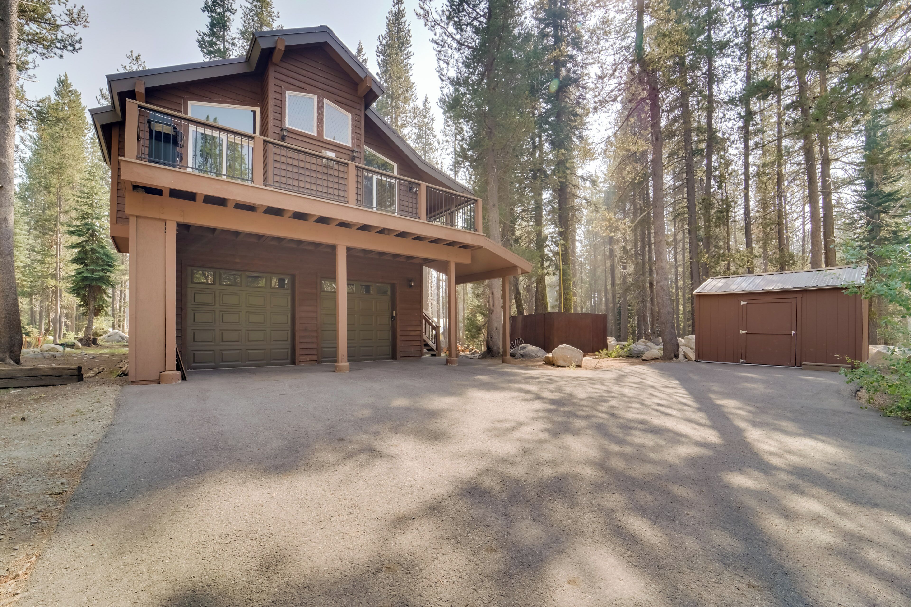 A two-story wooden cabin with a covered garage and a small shed, surrounded by tall trees.