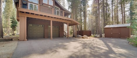 A two-story wooden cabin with a covered garage and a small shed, surrounded by tall trees.