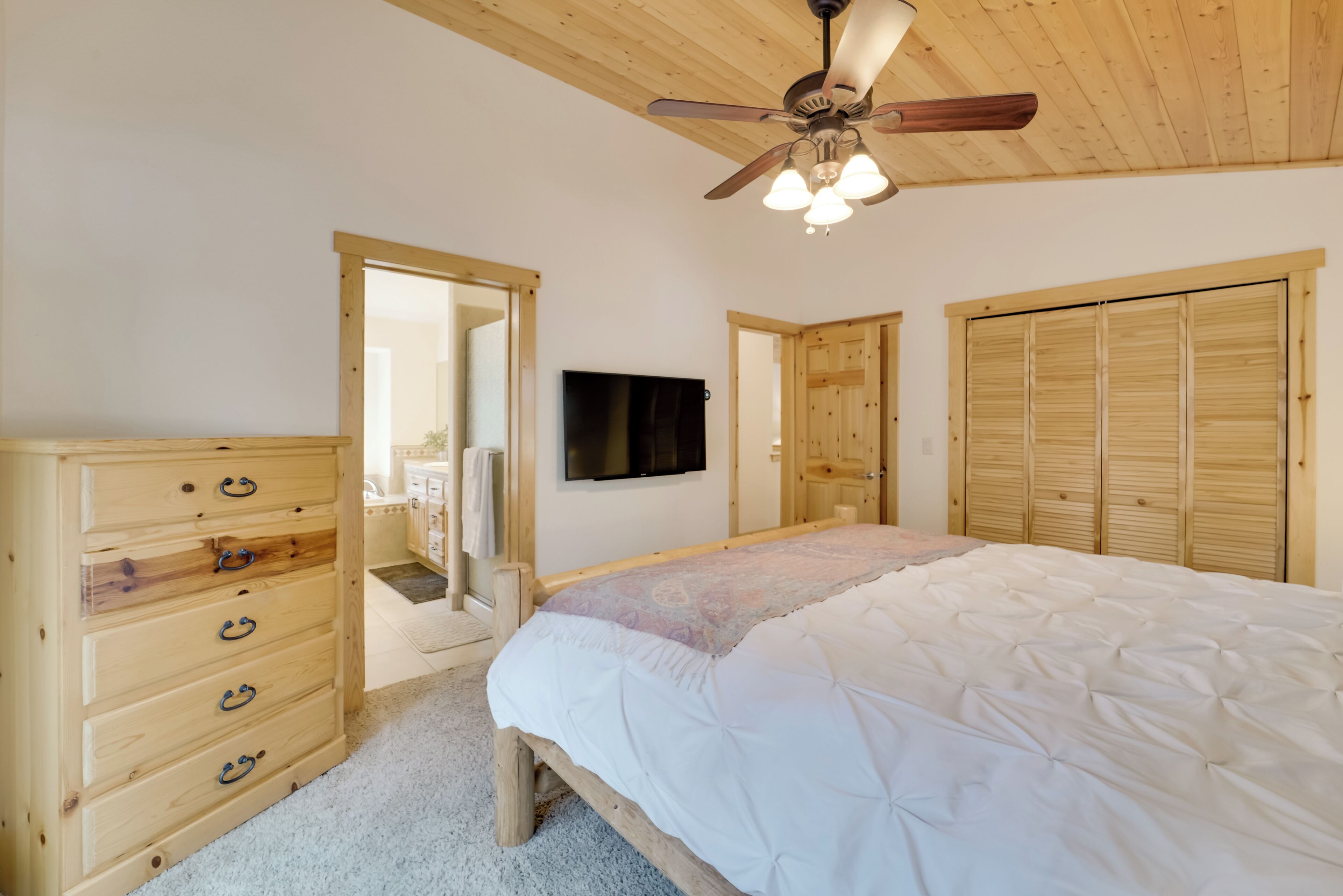 A bedroom with a wooden dresser, a bed with a white duvet, a ceiling fan, and wooden closets.