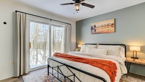 A bedroom with a bed, a ceiling fan, a window with curtains, a bedside table with a lamp, and a painting on the wall.