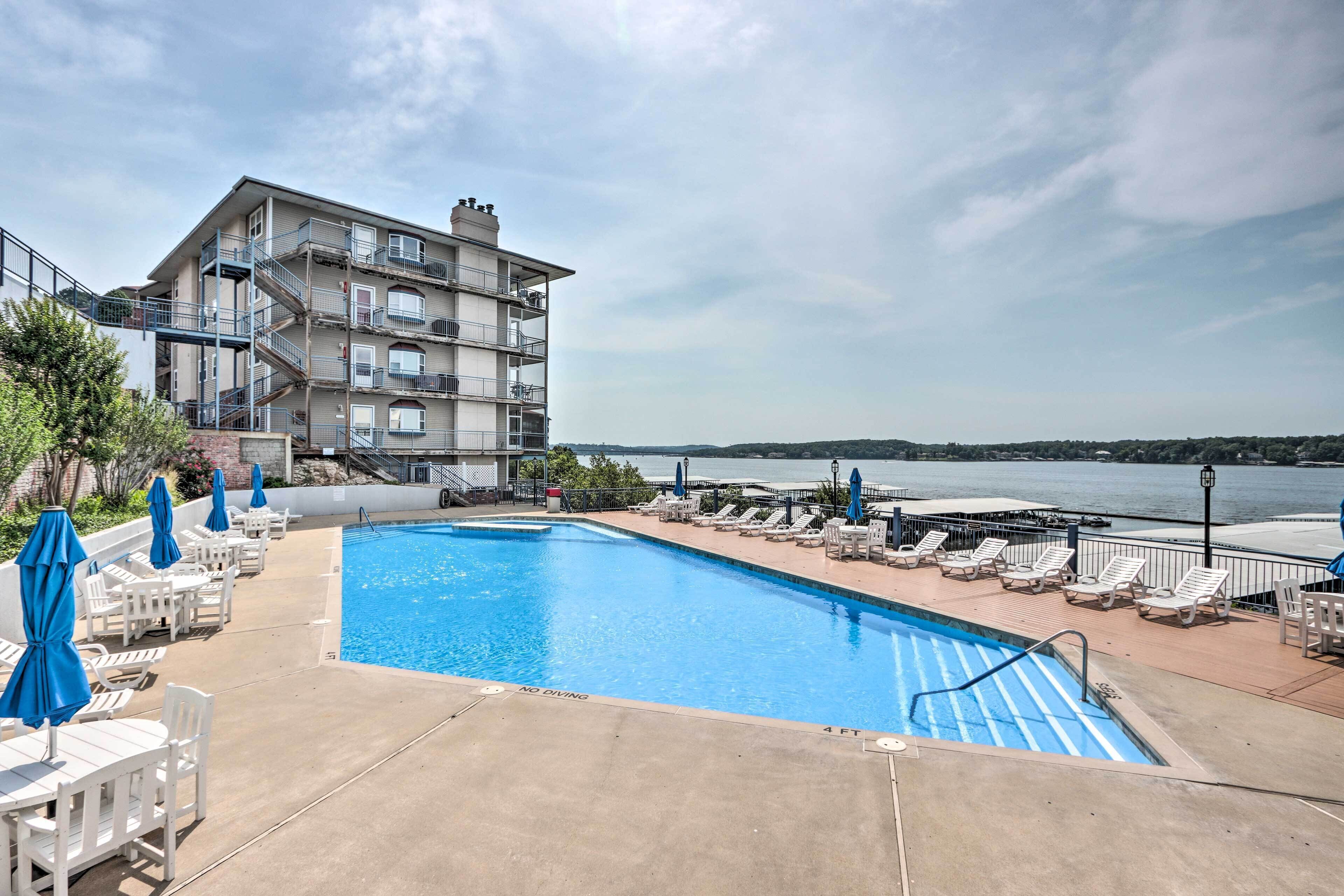 A swimming pool with lounge chairs and umbrellas, a building with multiple balconies, and a view of a body of water.