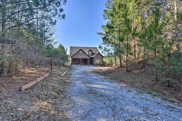 A house with a gravel driveway leading to a wooden door, surrounded by pine trees.