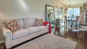 Una sala de estar con un sofá color beige, almohadones estampados, una manta roja, un área de comedor con mesa y sillas, y un espejo grande en la pared.