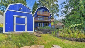Une maison à deux étages, avec une façade bleue, une terrasse en bois et un jardin où poussent différentes espèces de plantes.