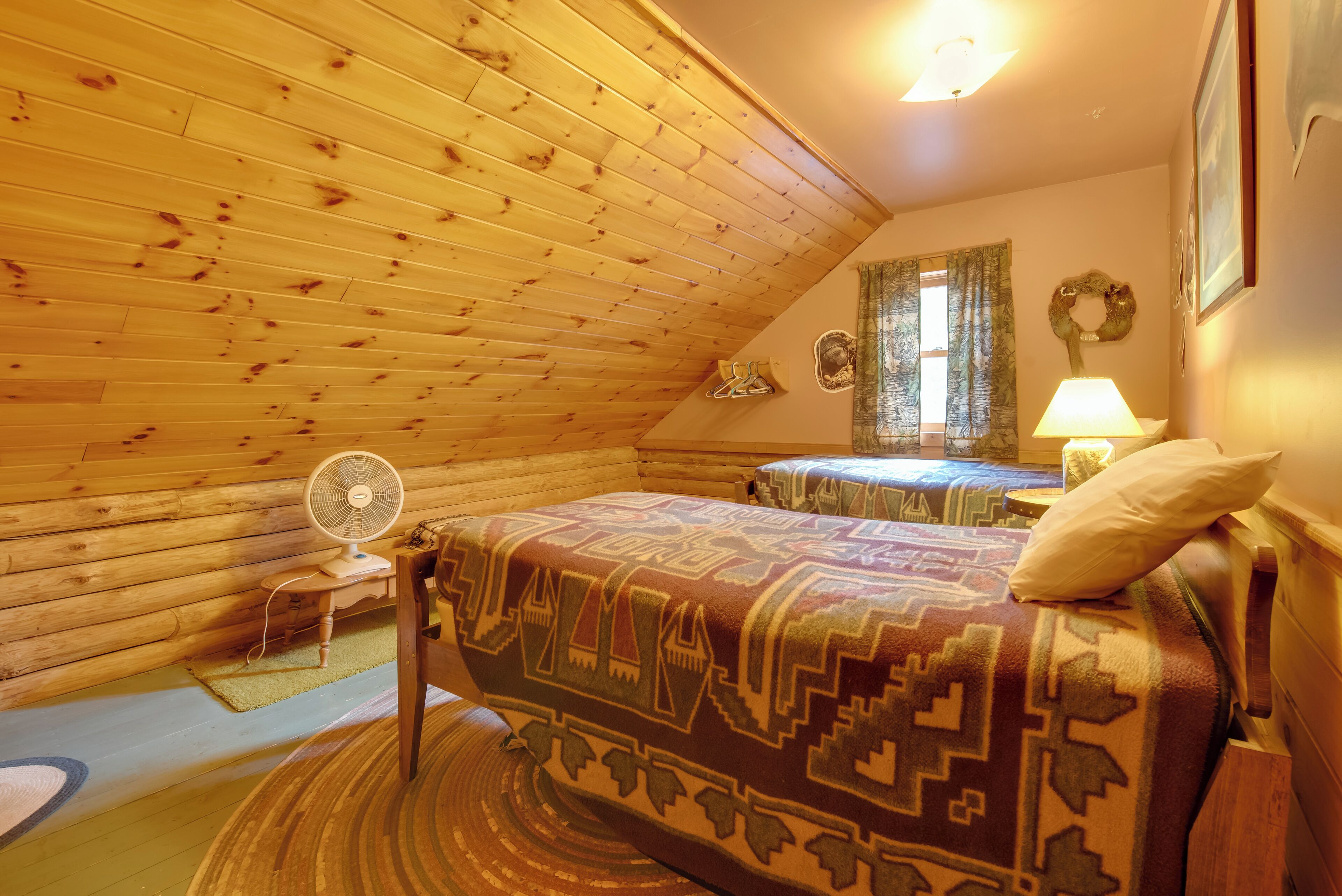 Ein gemütliches Schlafzimmer im Dachgeschoss mit Holzbalken an der Decke, einem Bett mit gemustertem Überwurf, einem Nachttisch mit Lampe und einem Ventilator auf dem Boden.