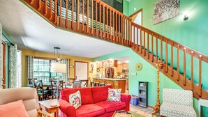 A spacious living room with a red sofa, a green ottoman, and a patterned armchair. A wooden staircase with a railing leads to an upper level.