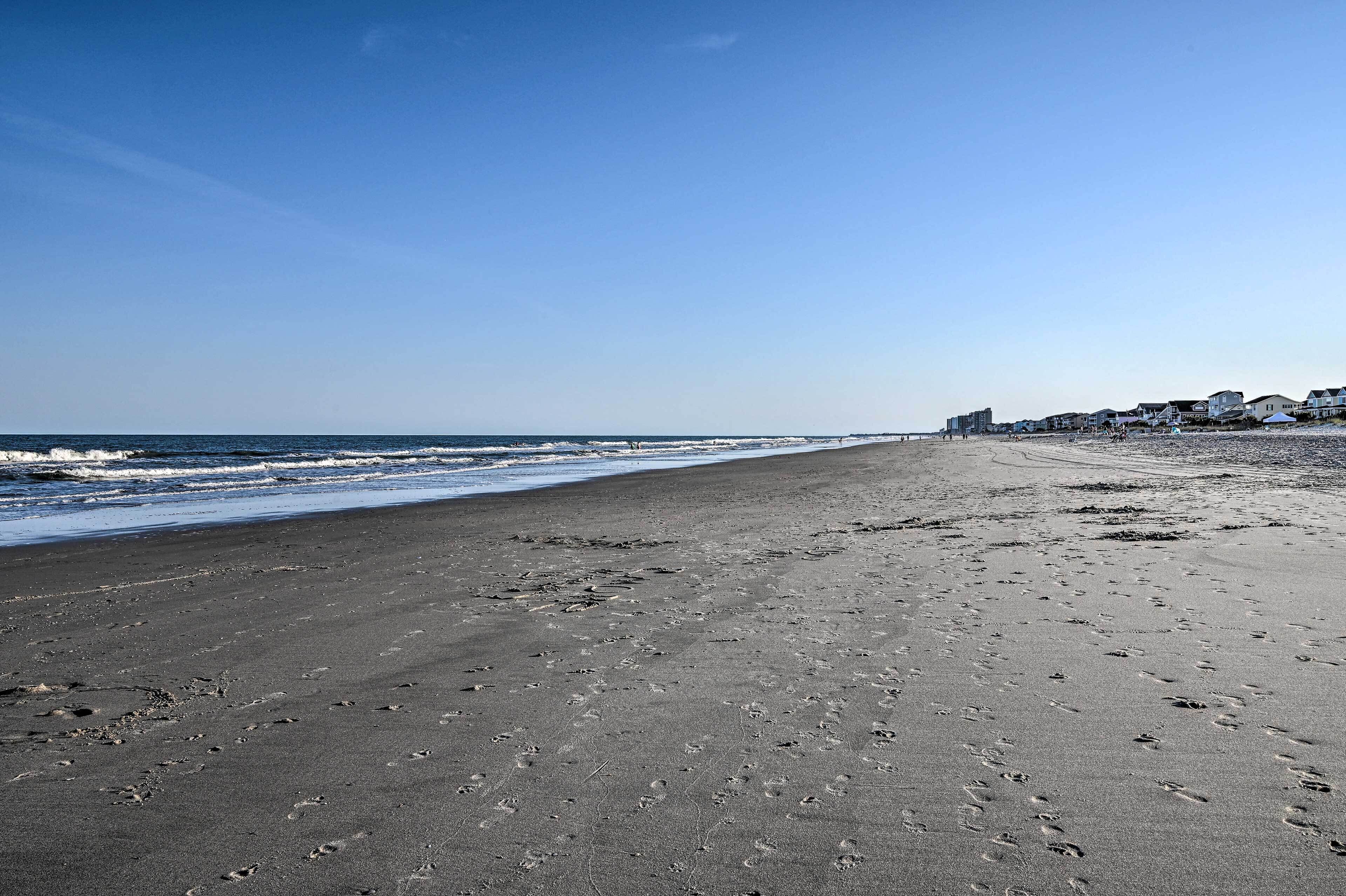 Une vaste plage parsemée d’empreintes, un ciel dégagé et des bâtiments visibles à l’arrière-plan.