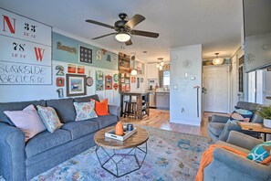 A living room with a grey sofa, a coffee table, and a ceiling fan. There are wall decorations and a kitchen in the background.