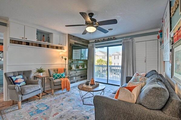 A cozy living room with a grey sofa, armchairs, a coffee table, and a ceiling fan. There is a large window with a view of houses and a balcony.
