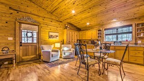 Interior de una cabaña de madera con comedor, dos sillones, una mesa y una cocina con armarios de madera y un ventanal.