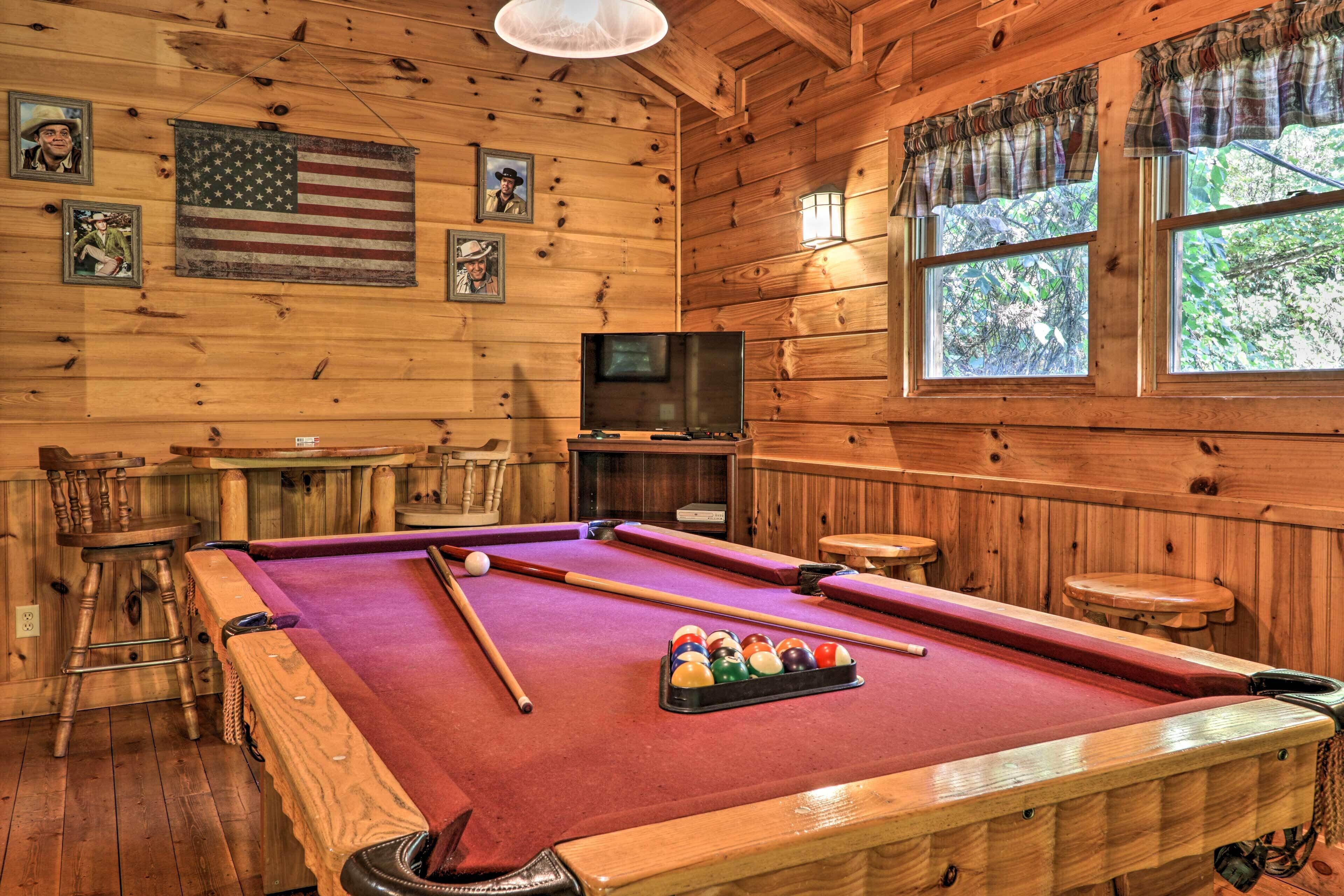 A wooden pool table with a pool cue and balls, a television, and framed pictures on the wall.