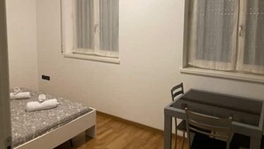 A small, single-room apartment with a bed, a table, and two windows with curtains. - house-parking-wifi-adige muse (Trento)