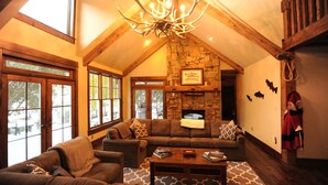 A cozy living room with a stone fireplace, large windows, and a chandelier. - High End Lodging for Medium to Large Groups in the Canyon of the Missouri River! (Craig)