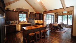 A spacious kitchen with wooden cabinets, a large island with bar stools, and a dining area with a stone wall and large windows. - High End Lodging for Medium to Large Groups in the Canyon of the Missouri River! (Craig)