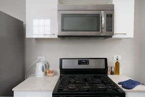 Une cuisine équipée d’un four à micro-ondes en acier inoxydable placé au-dessus d’une cuisinière à gaz, d’une bouilloire et d’un récipient contenant des capsules de café.