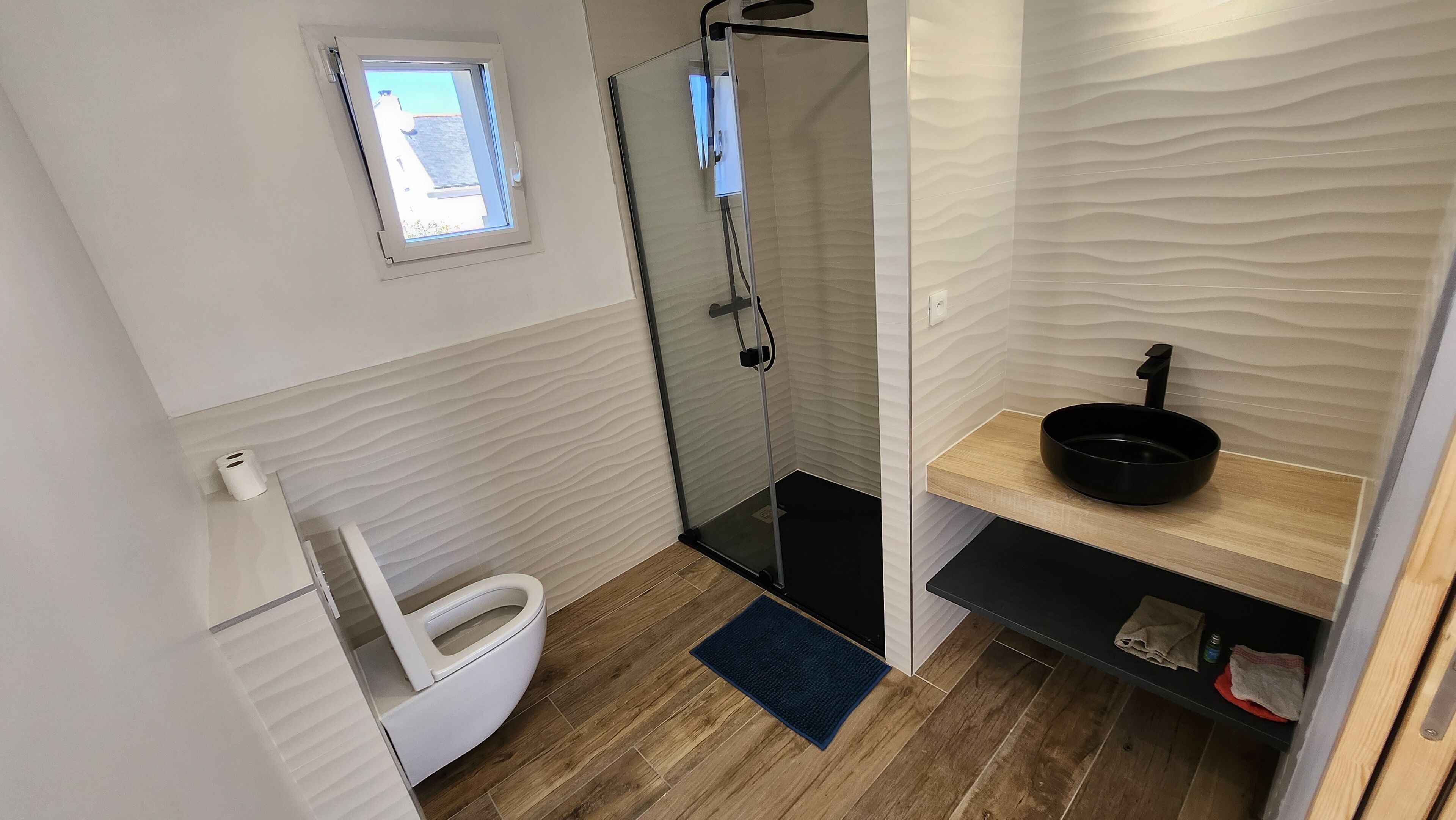 Een compacte badkamer met toilet, een glazen douchecabine, een wastafel met een zwarte wasbak en een klein raam boven het toilet.
