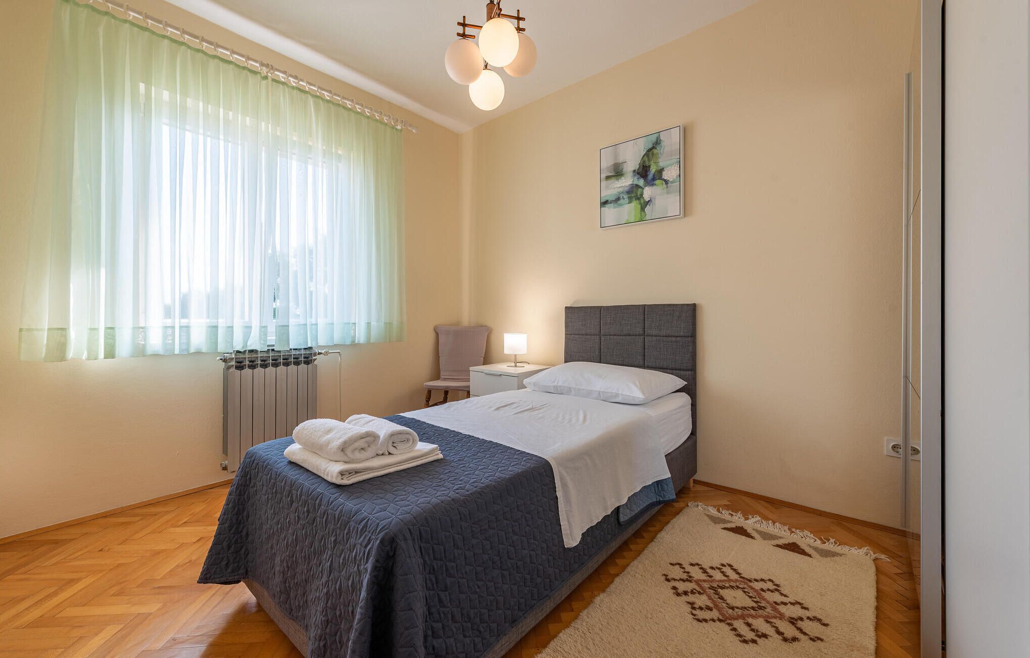 A hotel room with a bed, a nightstand, a lamp, a radiator, a window with sheer curtains, a painting on the wall, and a patterned rug.