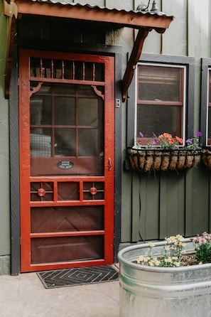 Une porte rouge avec un panneau de verre, un paillasson en mĂ©tal et une fenĂȘtre ornĂ©e dâun pot de fleurs suspendu.