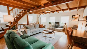 A spacious living room with a green sofa, wooden ceiling beams, a staircase, and a dining area with a wooden table and chairs.