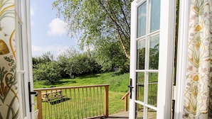 An open door to a deck with a wooden railing, a picnic table, and a grassy area with trees in the background.