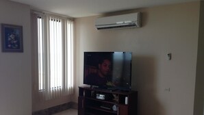 A living room with a flat-screen TV on a wooden stand, a ceiling-mounted air conditioner, and a window with vertical blinds. - Departamento en Manta Frente al mar (Manta)