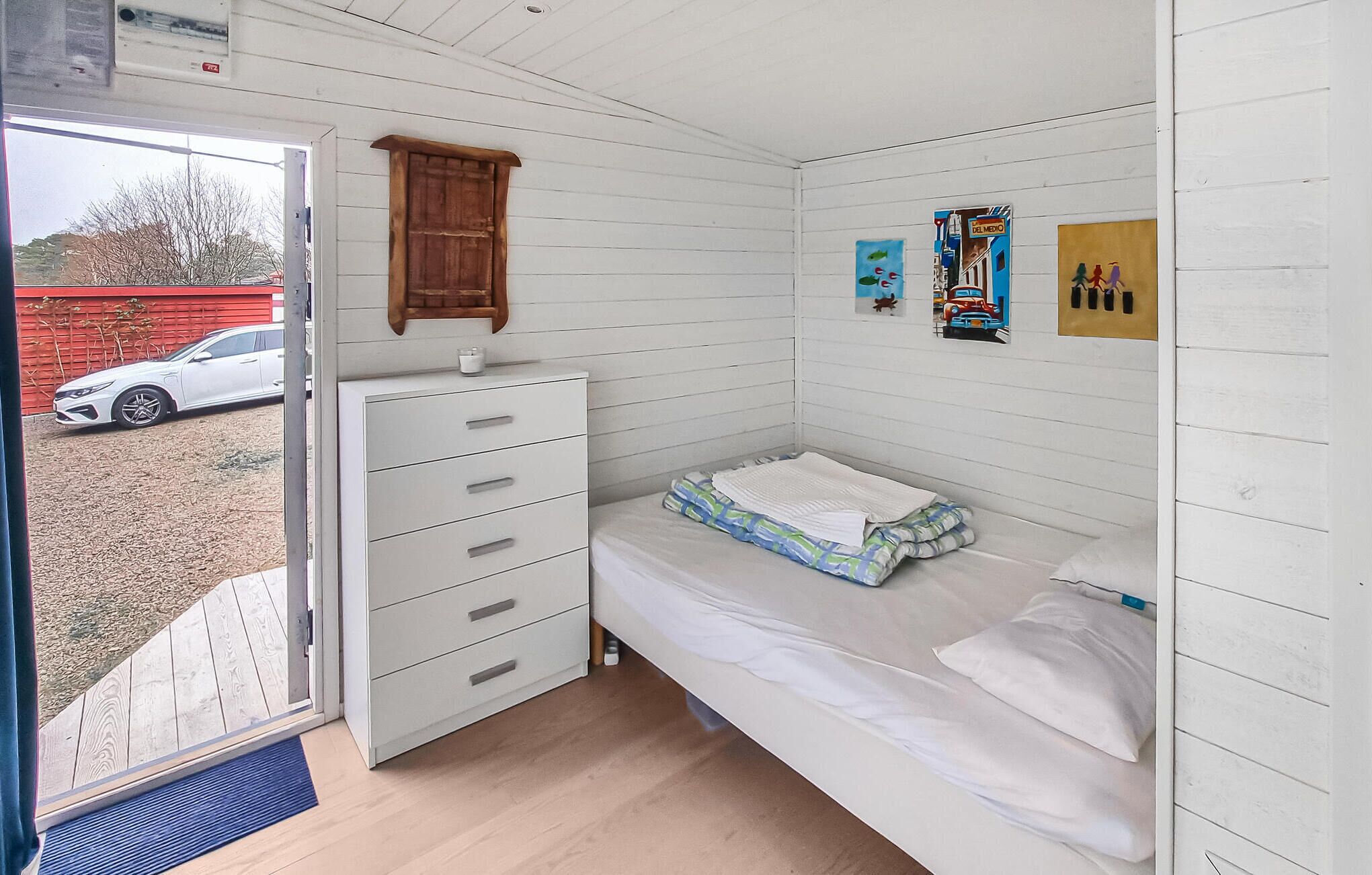 A small, white-paneled room with a bed, a dresser, and a door leading outside.