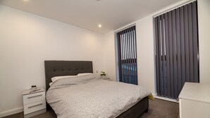 Une chambre à coucher comprenant un lit, une table de chevet, une commode et une fenêtre avec des stores.