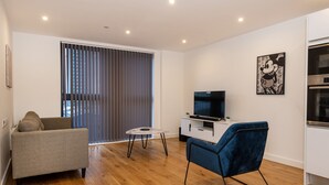 Un salon moderne avec un canapé gris, un fauteuil bleu, une petite table blanche et un téléviseur posé sur un meuble blanc.