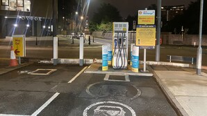 Un espace de stationnement nocturne équipé de bornes de recharge pour véhicules électriques, avec un panneau « Shell Recharge » et un magasin « Toybox » en arrière-plan.