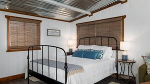 A bedroom with a metal bed frame, white bedding, two bedside tables with lamps, a green chair, and a wooden floor. - Welcome to the Ladybug Walk DT and Blanco River (Wimberley)