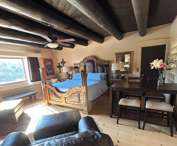 Une chambre à coucher avec un lit à baldaquin, un bureau avec une chaise, une table à manger avec des chaises, un ventilateur de plafond et une fenêtre donnant sur les arbres.