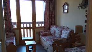 A cozy room with a wooden floor, a sofa, a small table, and a chair. There is a window with curtains, a lamp, and a small wall decoration. - MOUNTAIN-STYLE APARTMENT AT THE FOOT OF THE SLOPES SLEEPS 5 (La plagne - Belle-plagne)