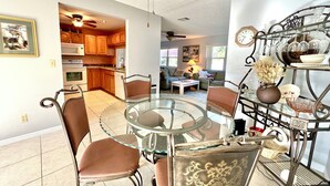 A dining area with a glass table and metal chairs, a kitchen in the background, and a living room with a sofa and lamp. - Boutique Stay: Near Beach,  PSP4, Billiard or PingPong, Fire Pit, Private Yard (New Port Richey)