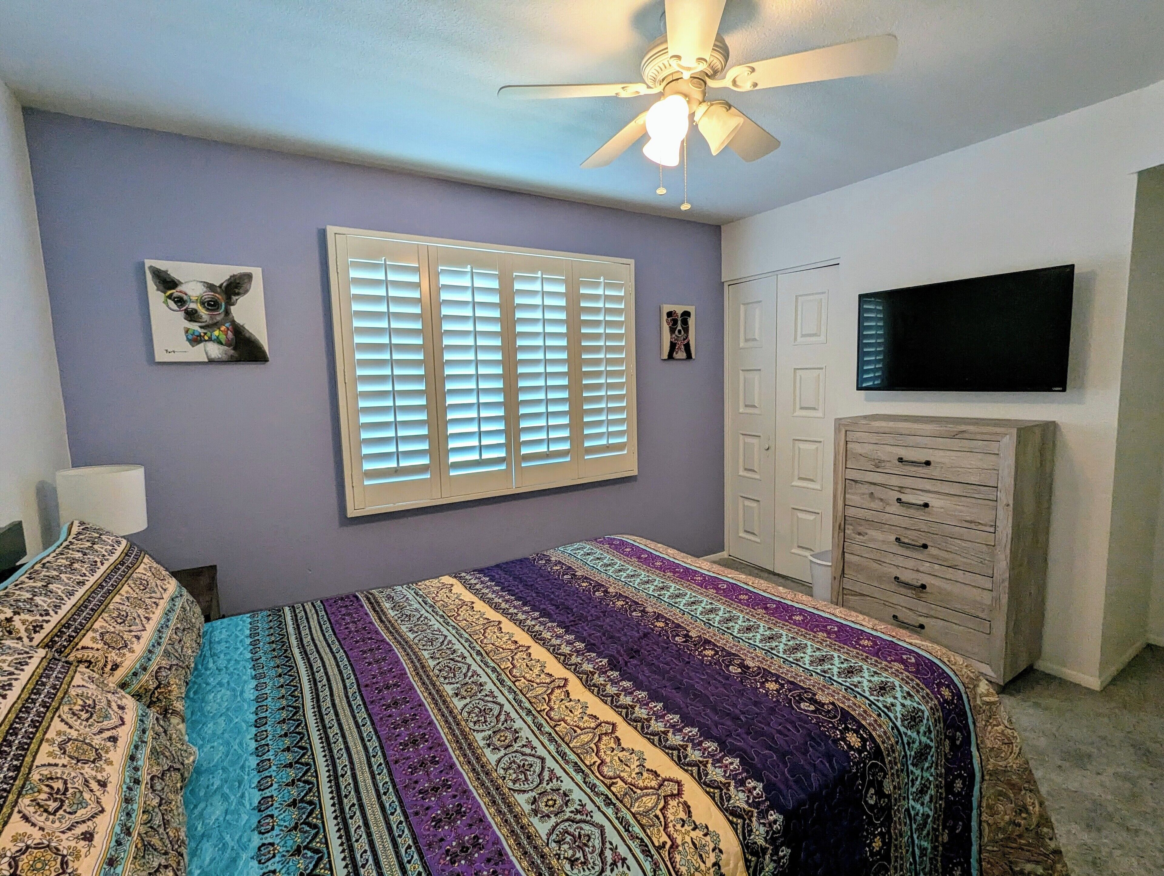 A bedroom with a bed, a ceiling fan, a window with blinds, a dresser, and a television on the wall.