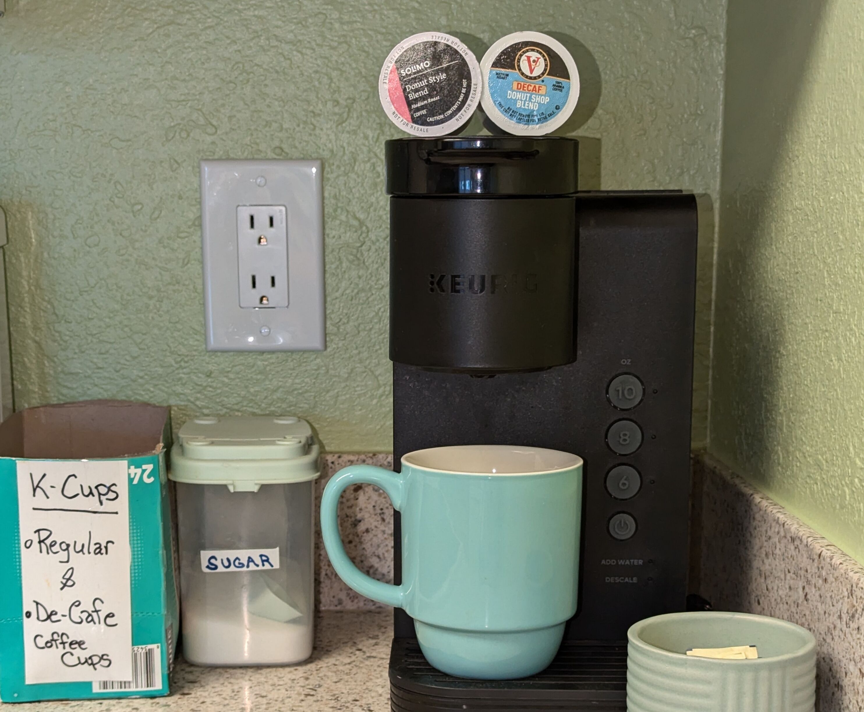 A Keurig coffee maker with two coffee pods, a K-Cups box, a sugar container, and a blue mug on a kitchen counter.