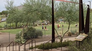 A park with a shaded area, a fenced dog area, and a pathway leading to a building with a sign.