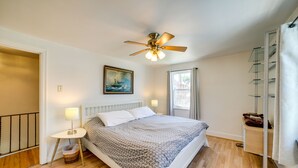 Een slaapkamer met een bed, een plafondventilator, een schilderij, een raam en een nachtkastje met een lamp.