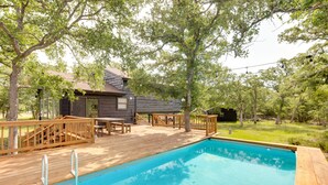 Une terrasse en bois avec une piscine, entourée d’arbres et d’un petit bâtiment dont l’extérieur est également en bois.