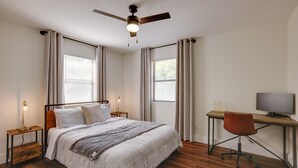Une chambre à coucher comprenant un lit, un bureau, une chaise, un ventilateur de plafond et deux fenêtres avec des rideaux.