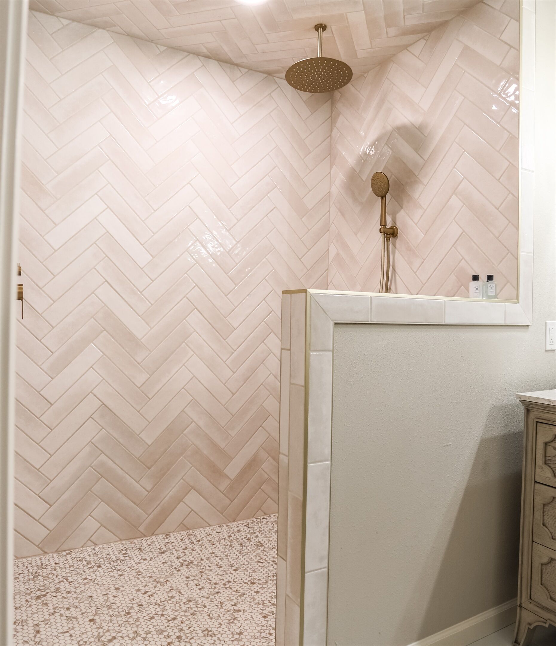 Une salle de bain avec un carrelage disposé en motif chevron sur les murs et le sol, une douche dotée d’un pommeau de douche doré et un petit miroir au-dessus du lavabo.