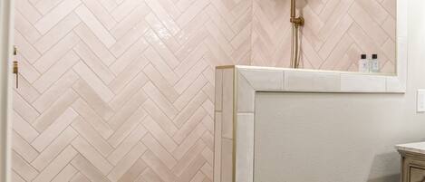 Une salle de bain avec un carrelage disposé en motif chevron sur les murs et le sol, une douche dotée d’un pommeau de douche doré et un petit miroir au-dessus du lavabo.