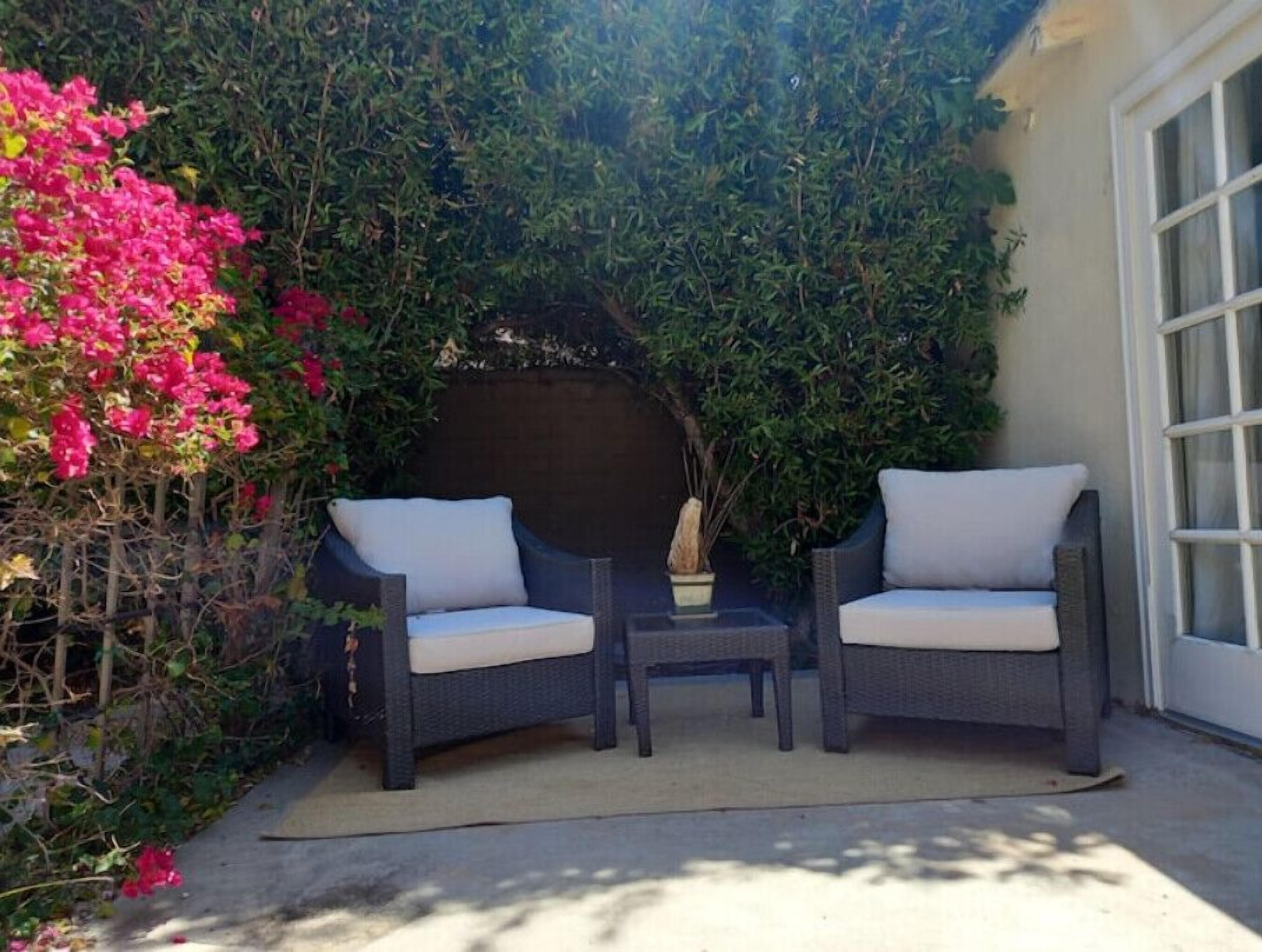 A patio with two wicker chairs and a small table, surrounded by lush greenery and pink flowers.