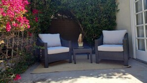 A patio with two wicker chairs and a small table, surrounded by lush greenery and pink flowers.