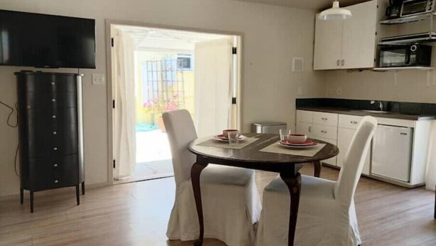 A modern kitchen with white cabinets, a black countertop, and a microwave. There is a dining area with a table and chairs, and a television mounted on the wall.