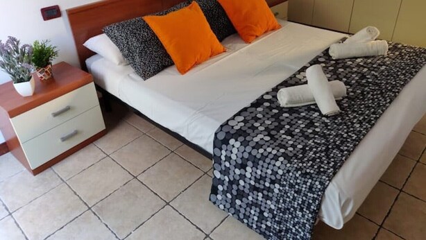 Una cama bien hecha con sábanas blancas, dos almohadas naranjas y una manta de estampado blanco y negro. Hay una mesita de noche con una planta y un cajón blanco.