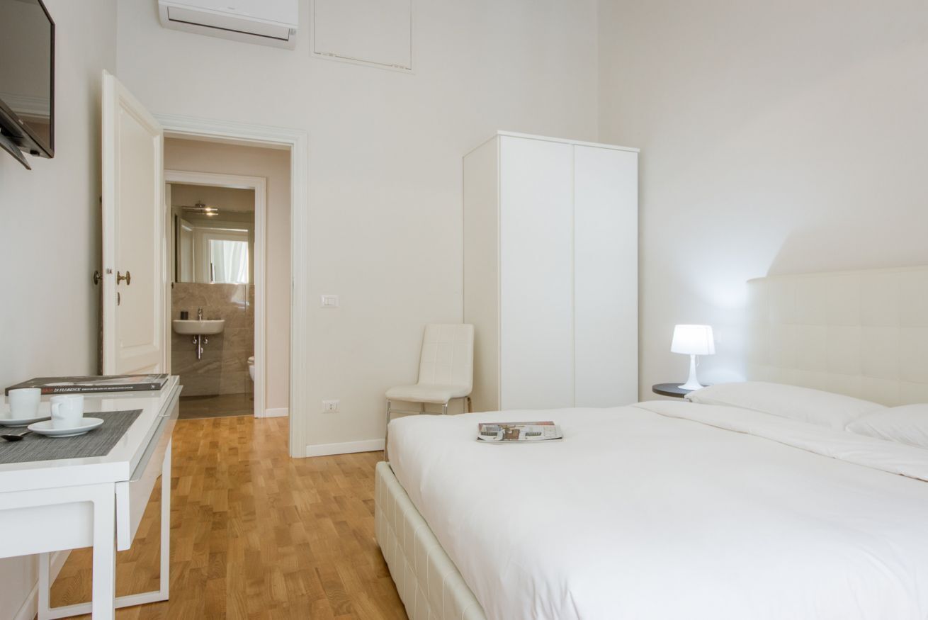 Une chambre d’hôtel comprenant un lit, une table de chevet, une chaise, une armoire, un bureau avec une chaise et une salle de bain visible à travers une porte ouverte.