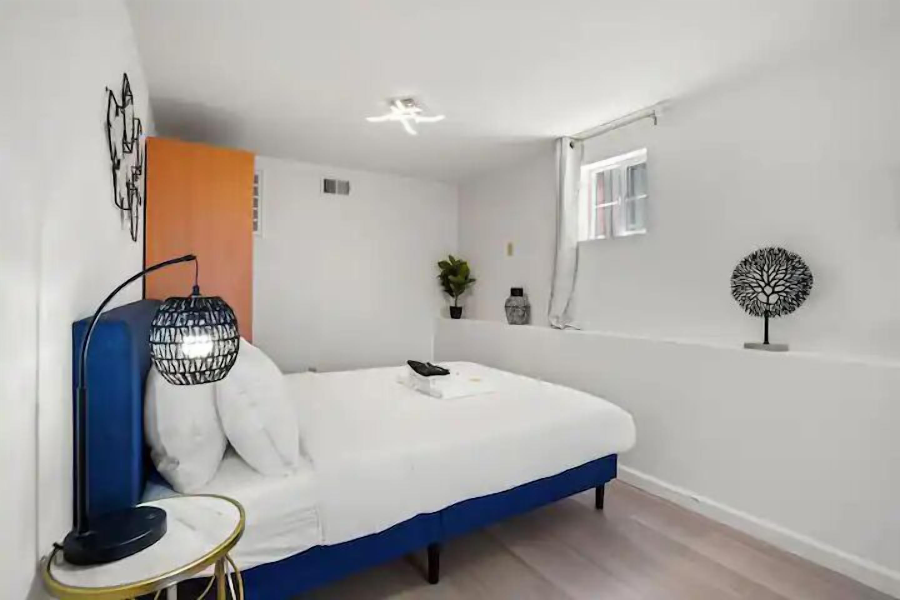 Un dormitorio con una cama individual, una mesita de noche, una lámpara, una planta en maceta y una estantería con objetos decorativos.