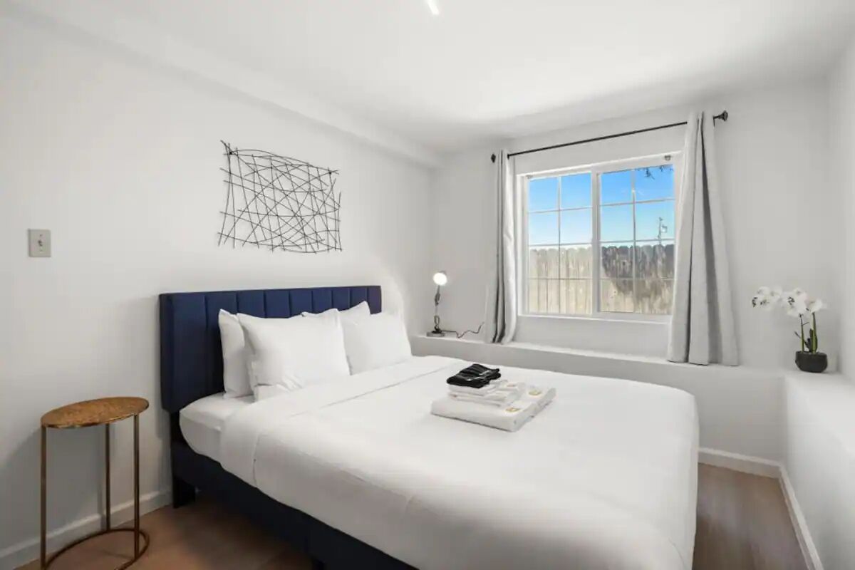 A modern hotel room with a large bed, a bedside table, a wall art piece, a window with curtains, and a view of the outdoors.